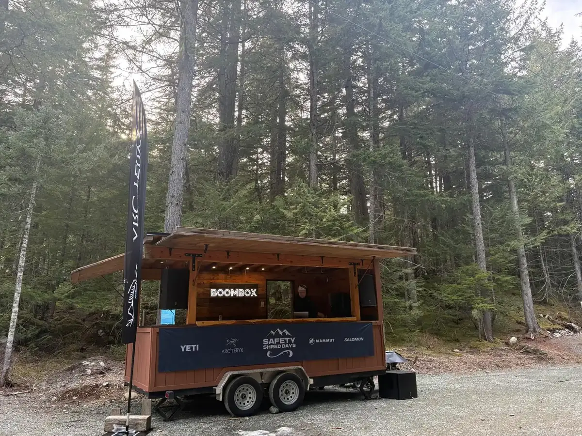 The branded Whistler Boombox trailer with an Arc'teryx feather flag and Safety Shred Days sponsor banner in a forest gravel lot